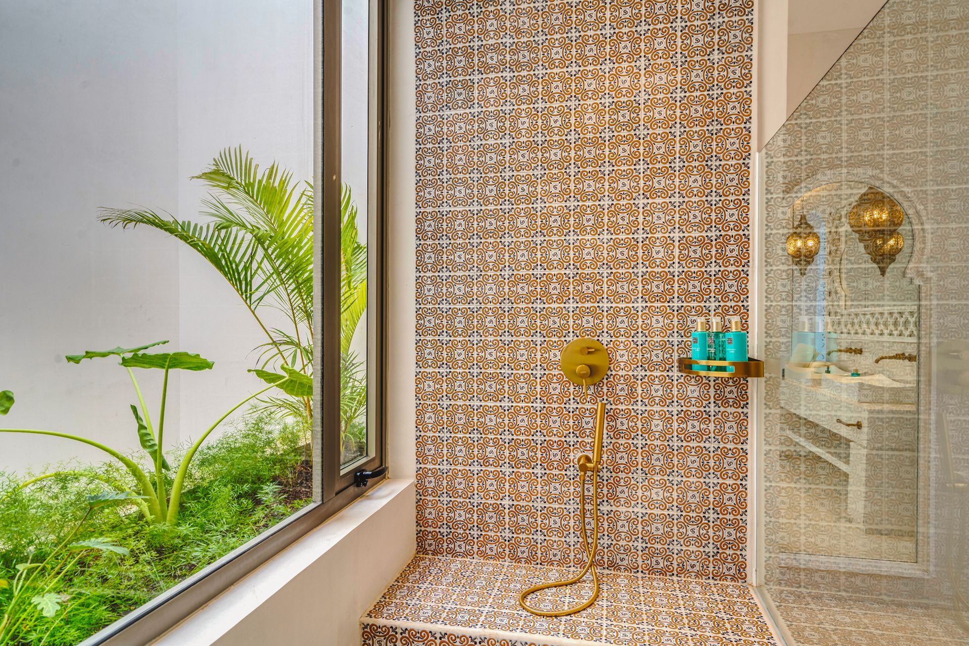 Ducha con azulejos estampados, accesorios dorados y una vista de una exuberante vegetación a través de una ventana.