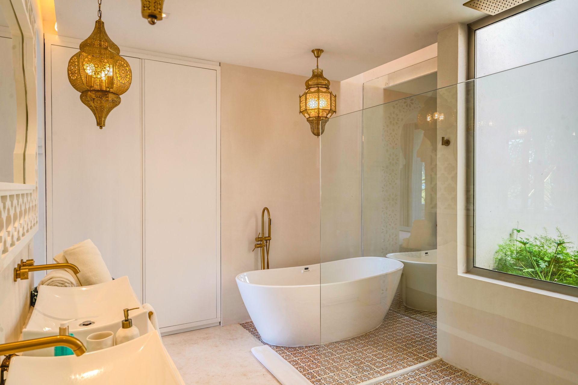 Elegante baño blanco con bañera independiente, dos lavabos, lámparas doradas decorativas y suelo de guijarros.