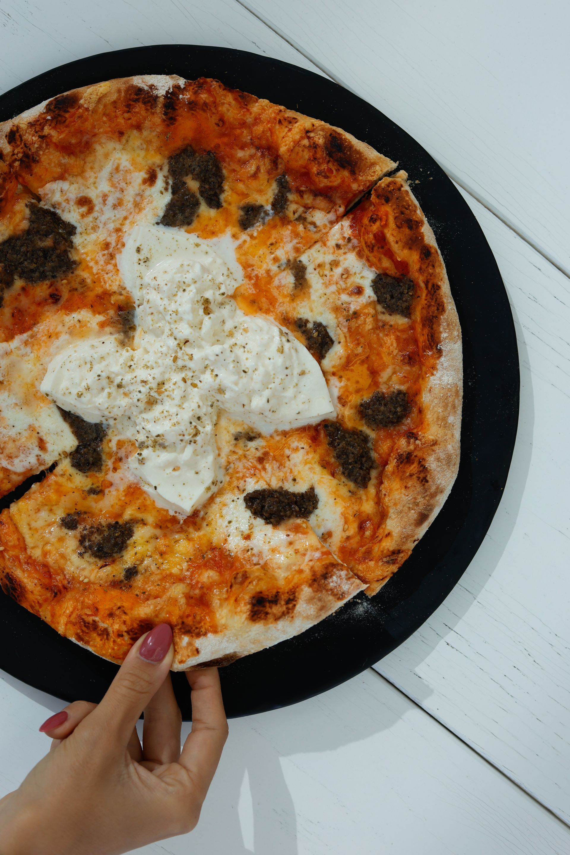 Pizza en un plato negro, con la mano extendida para tomar una rebanada. Queso blanco, salsa oscura y corteza dorada.