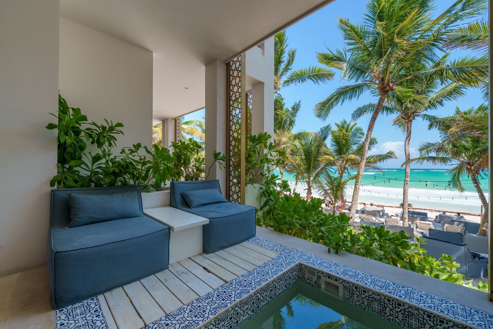 Balcón con asientos azules, pequeña piscina y vista a la playa con palmeras.