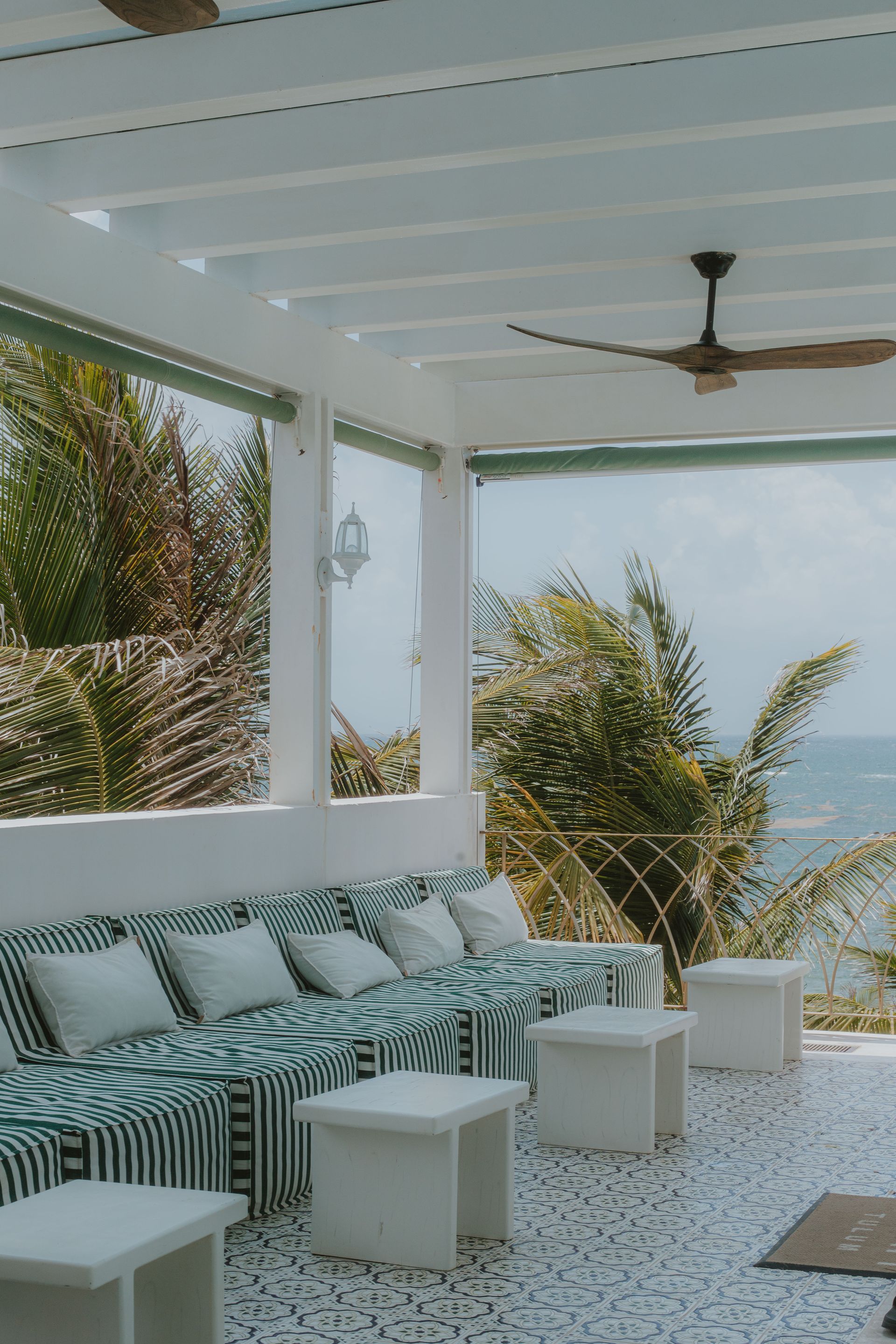 Una terraza blanca al aire libre con asientos acolchados de estampado verde y blanco, con vistas al océano y a las palmeras.