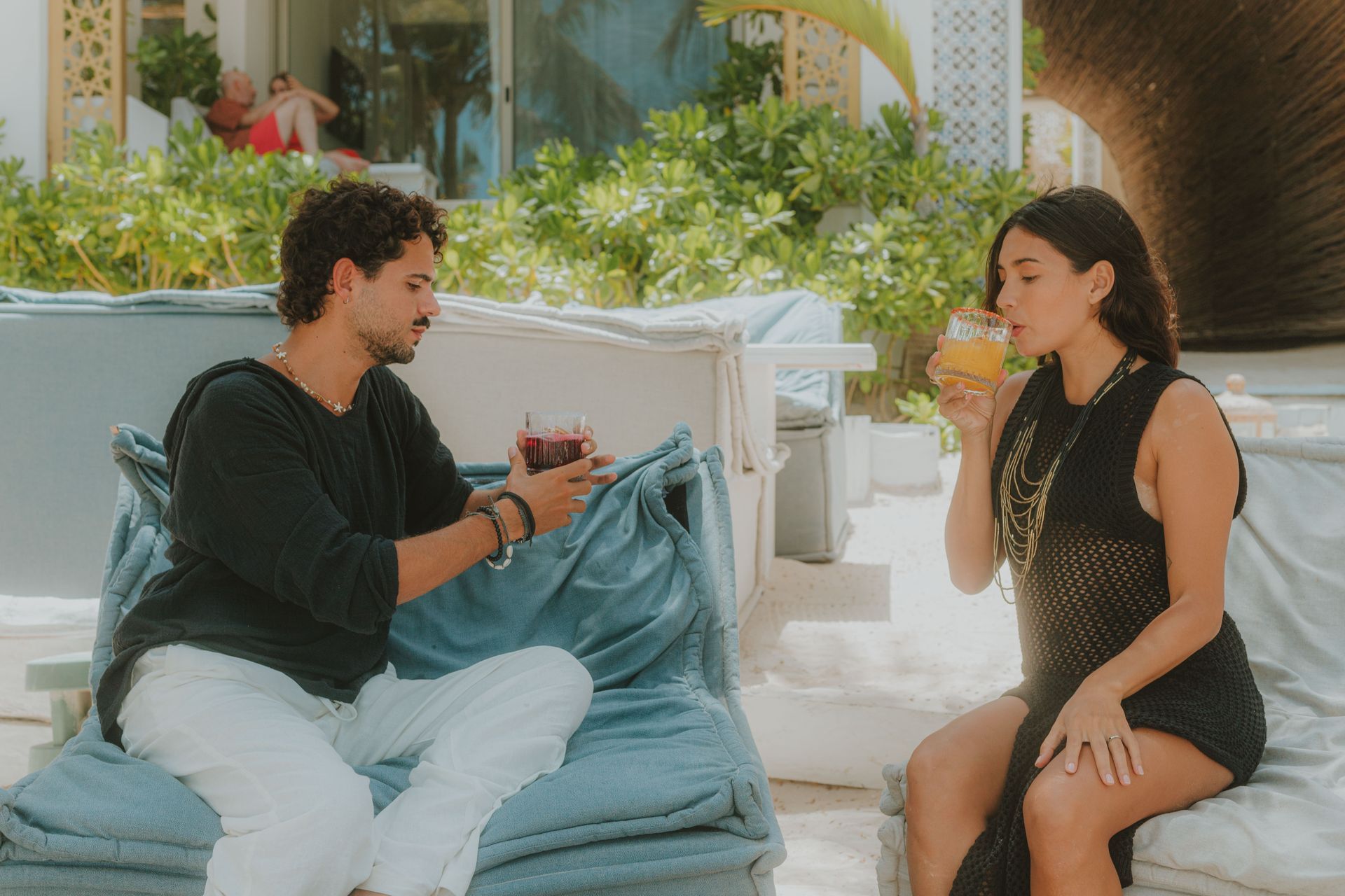 A man and woman drinking cocktails on a beach. They are both in casual wear, relaxing in the sun.