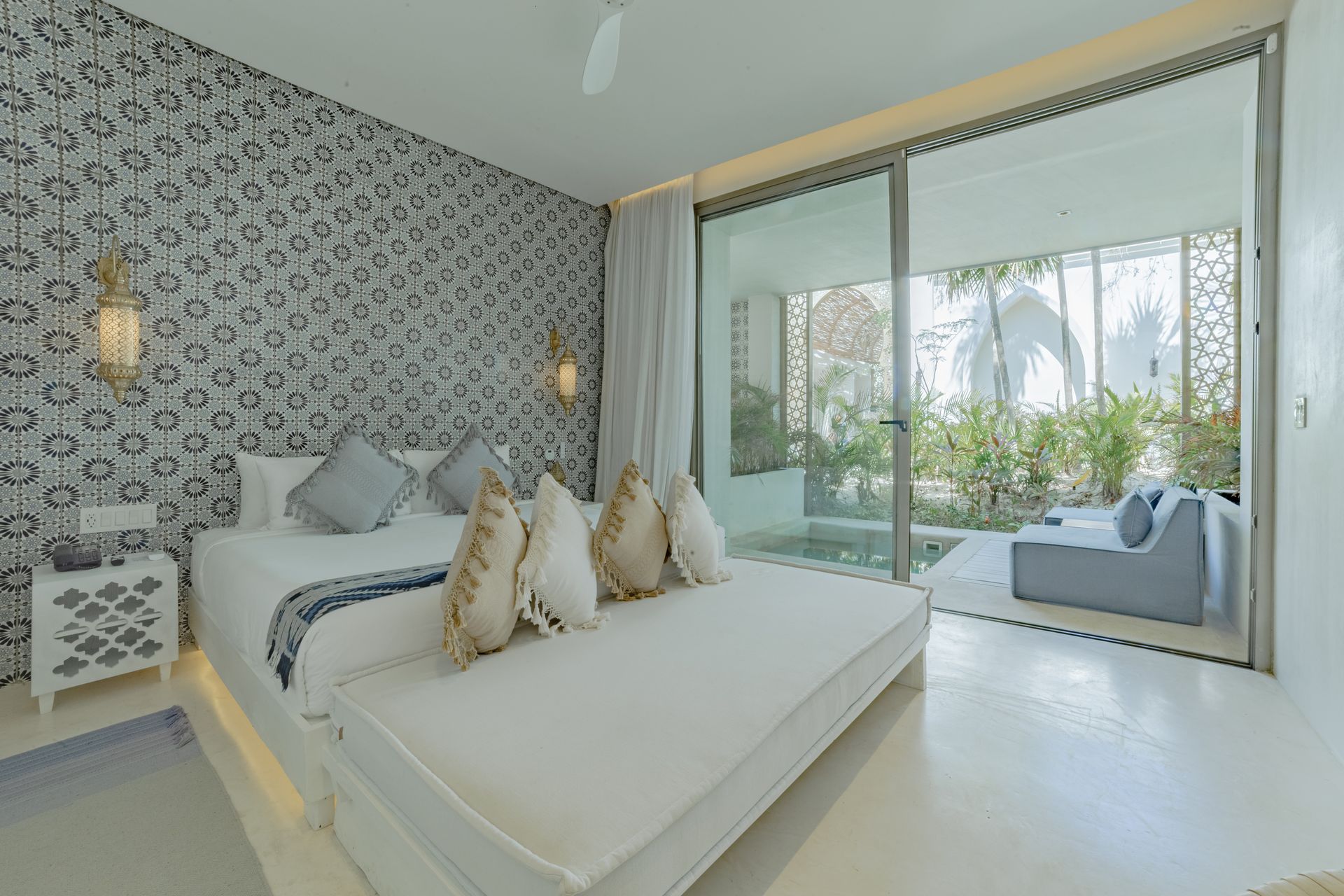 Dormitorio con cama blanca, papel tapiz estampado, puertas corredizas de vidrio que dan a un patio con piscina y asientos.