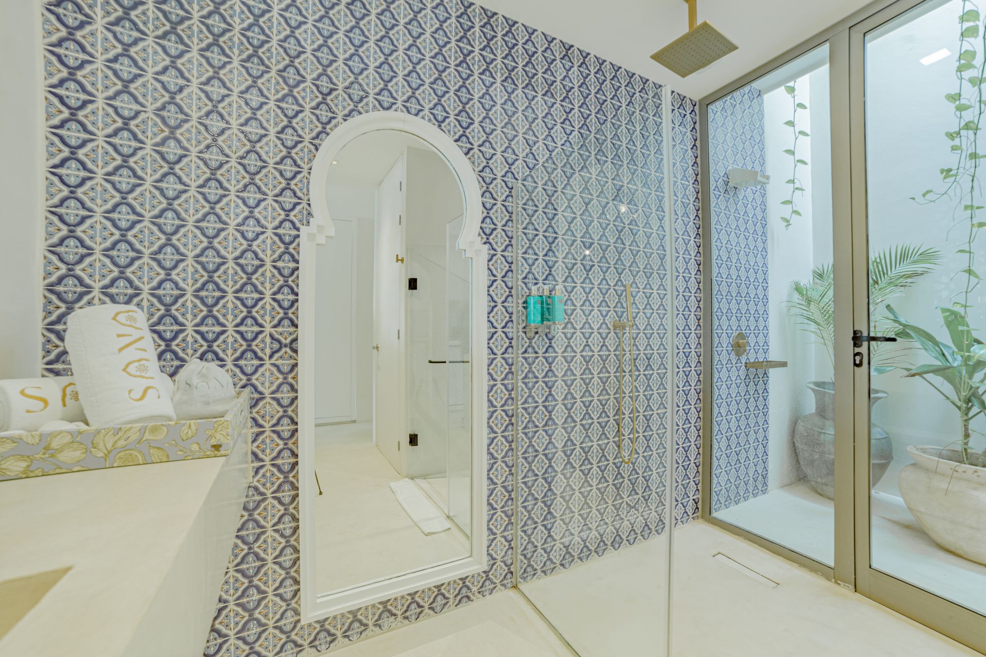 Baño con azulejos estampados en azul y blanco, ducha de cristal y vista al exterior con plantas.