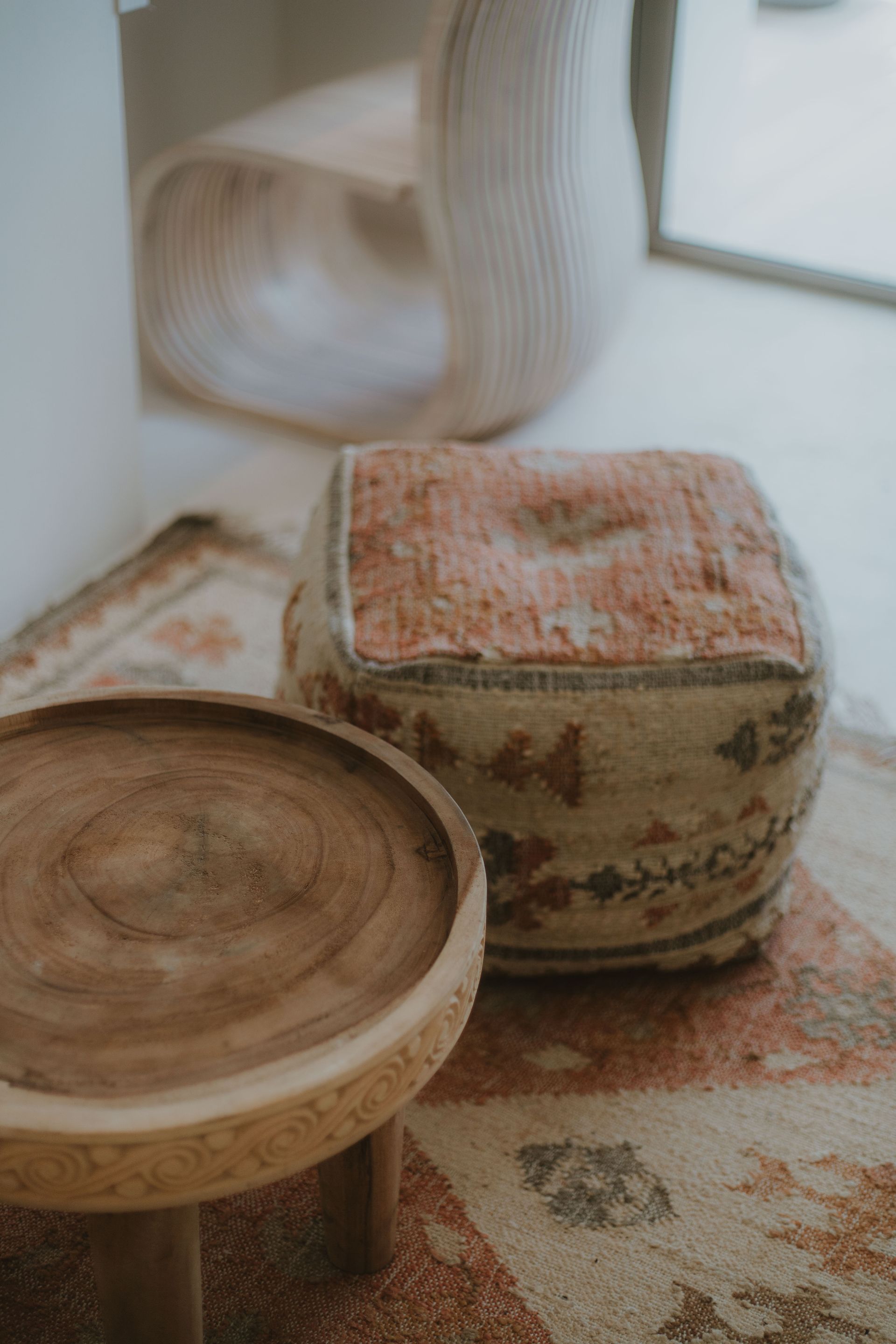 Mesa auxiliar de madera y puf estampado sobre una alfombra en una habitación luminosa.