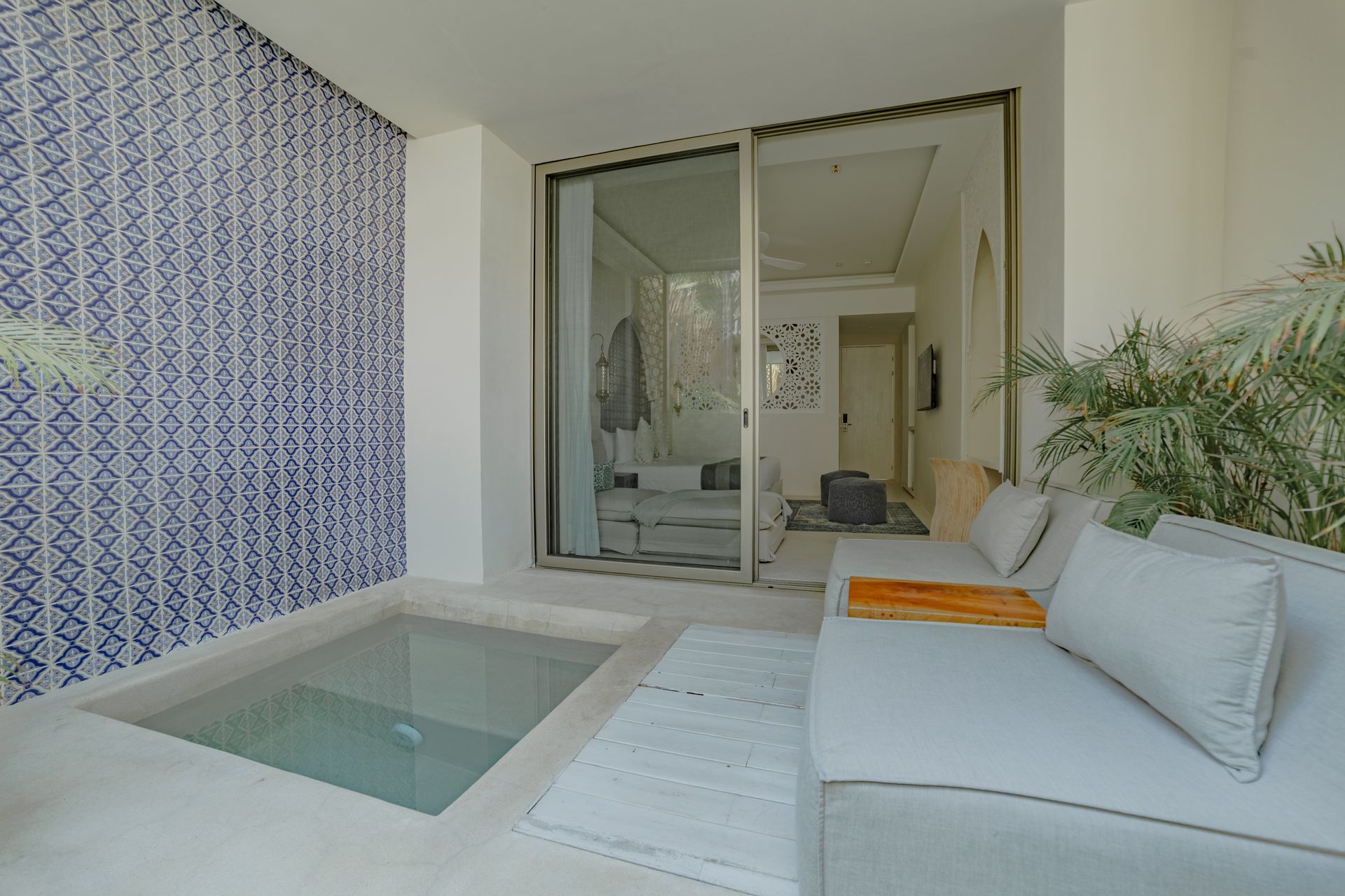 Espacio exterior con una pequeña piscina, pared de azulejos azules y un sofá blanco. Una puerta corrediza de vidrio da acceso a un dormitorio.