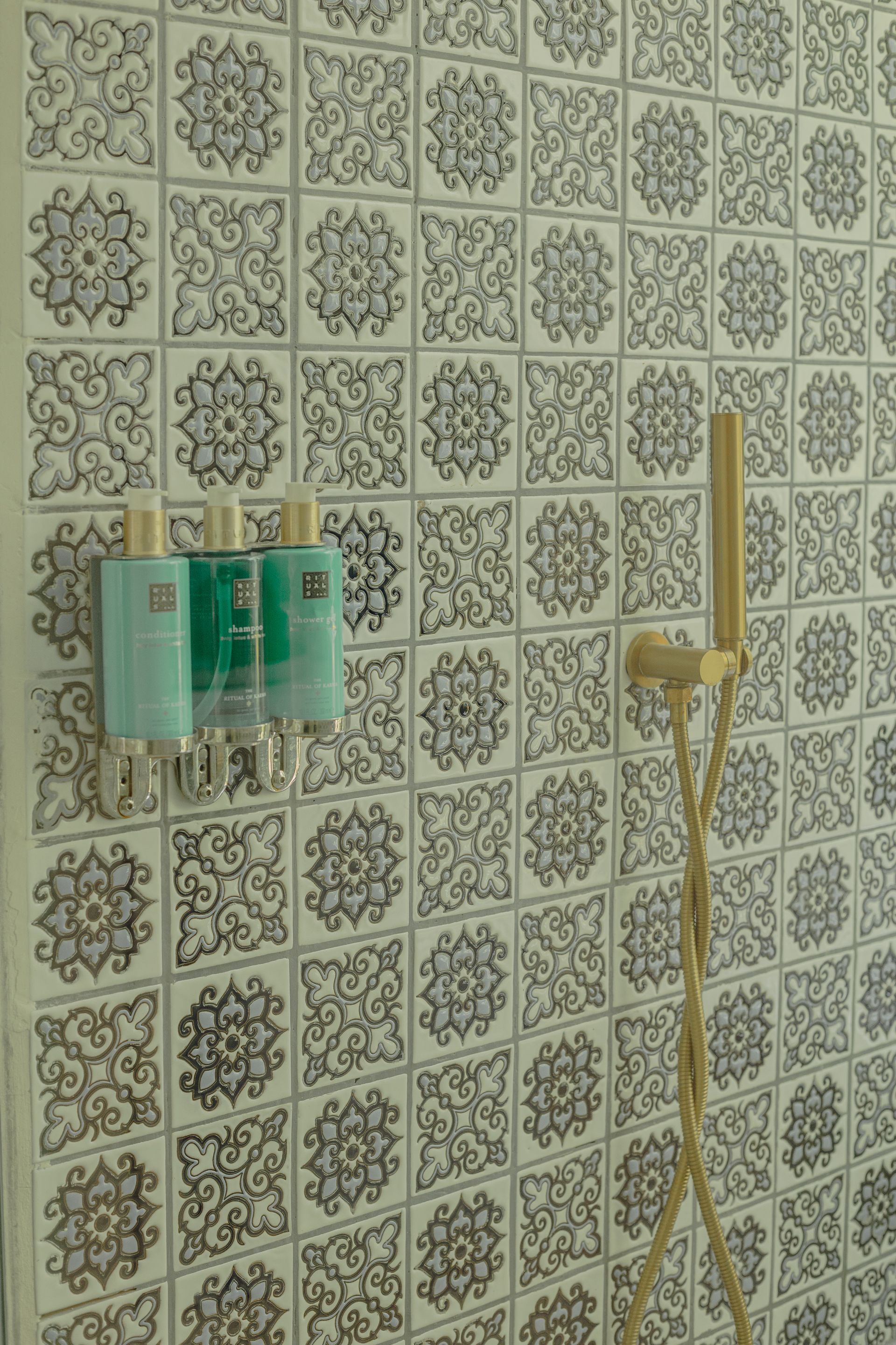 Ducha con pared de azulejos ornamentados, cabezal de ducha dorado y botellas de champú color verde azulado.