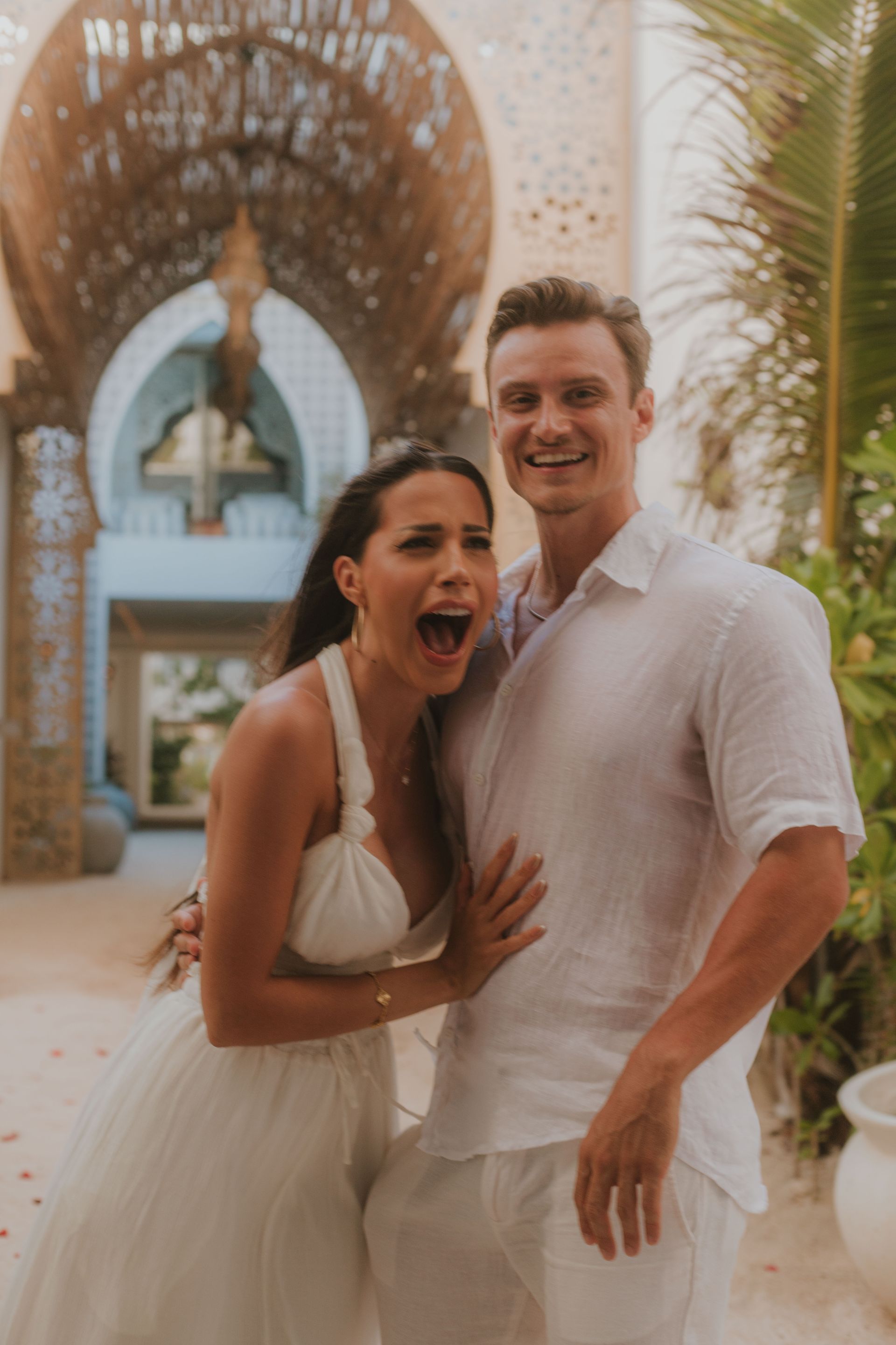 Couple in white clothing; woman with open mouth and joyful expression, man smiles, in front of ornate archway.
