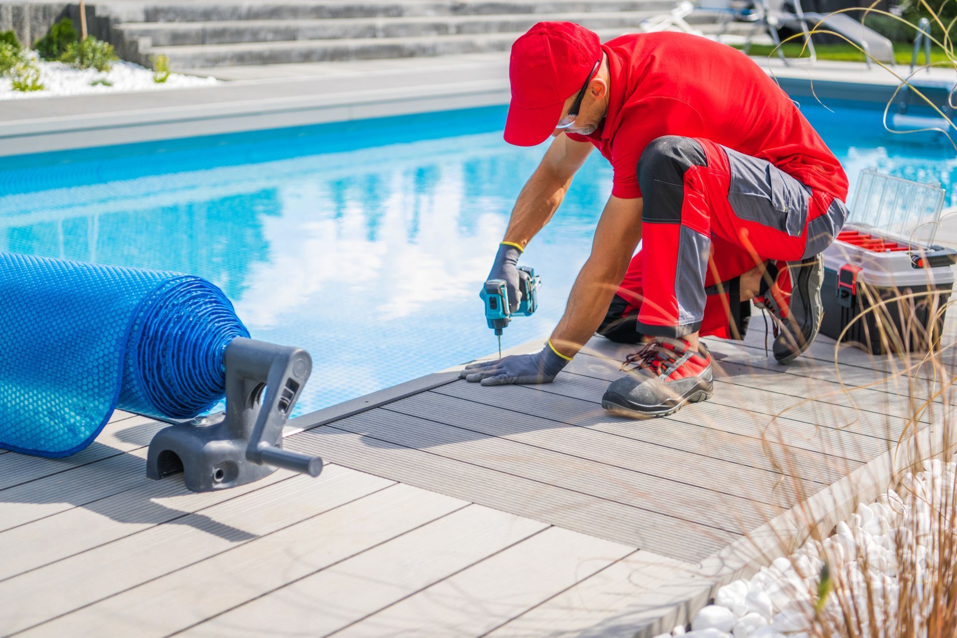 Man in red outfit installing pool cover near a pool, using a power drill.