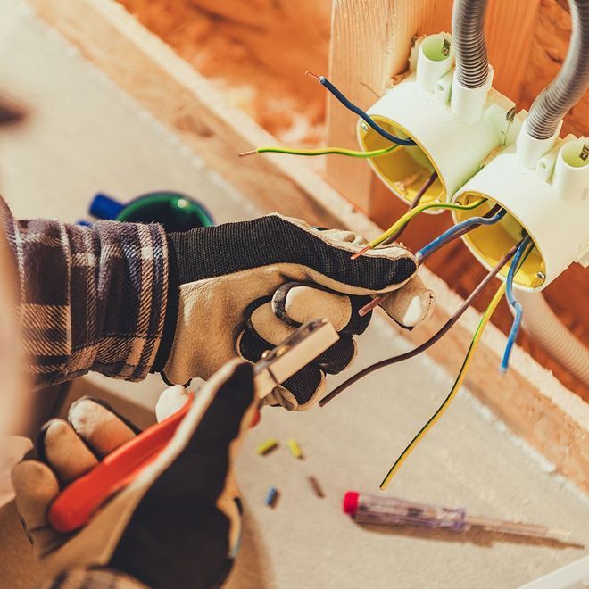 Electrician wiring electrical box, cutting wires with pliers, wearing gloves in a wooden frame setting.