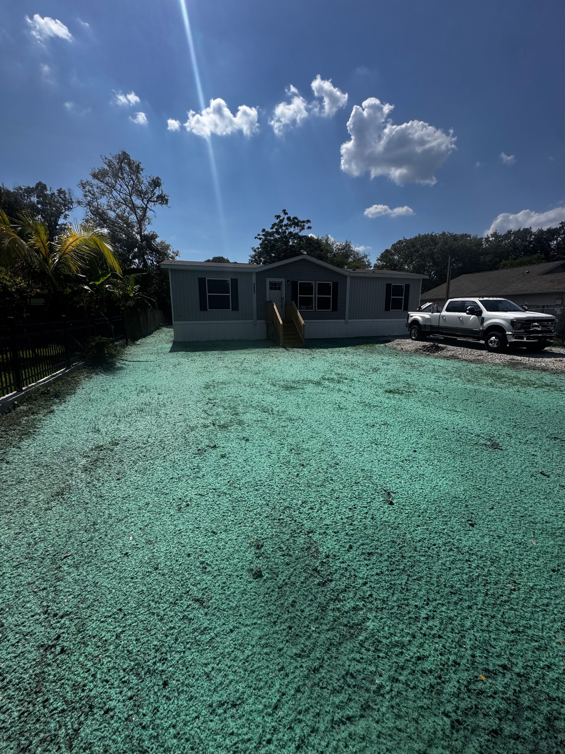 Mobile home with teal gravel yard; white truck parked in driveway, sunny day.