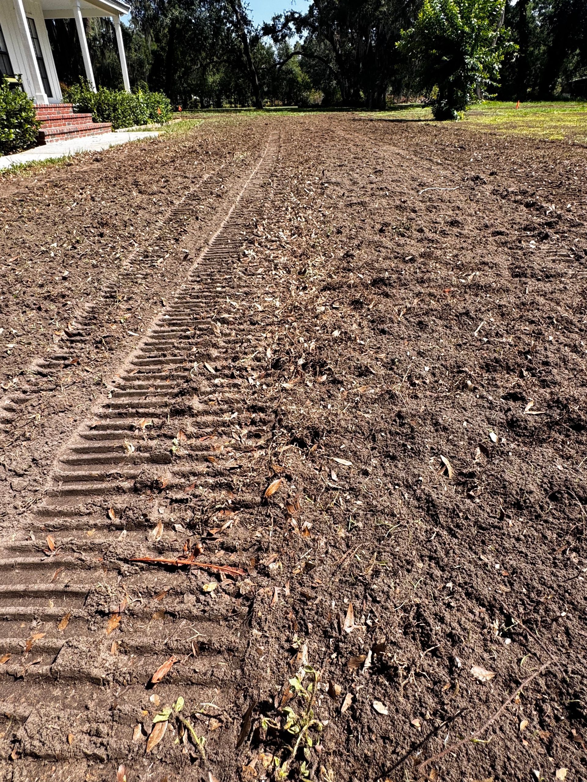 Tilled brown soil in a yard, with rows and tracks from machinery, near a house with a porch.