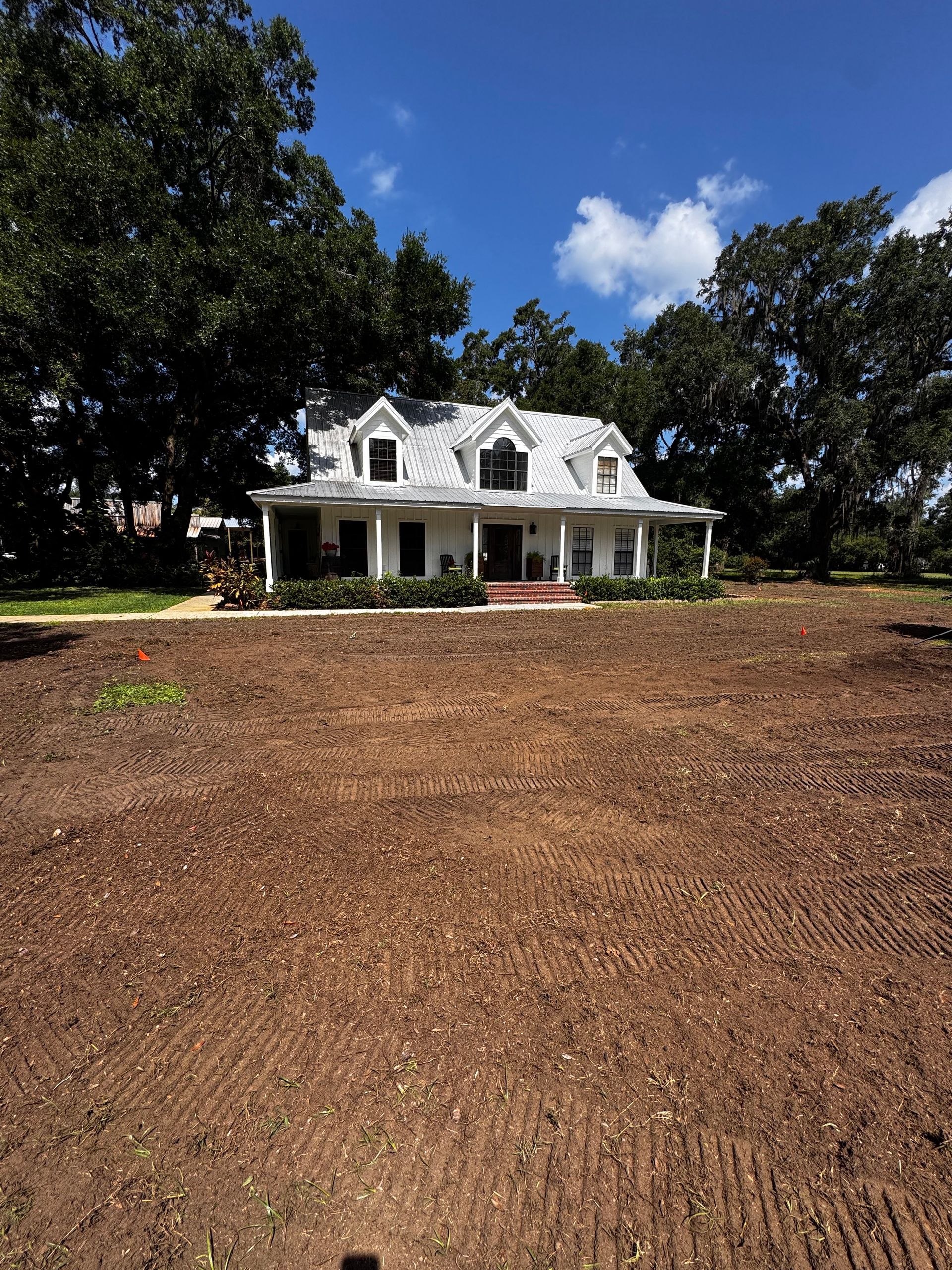 White house with gray roof, surrounded by trees, on a brown, barren yard under a blue sky.