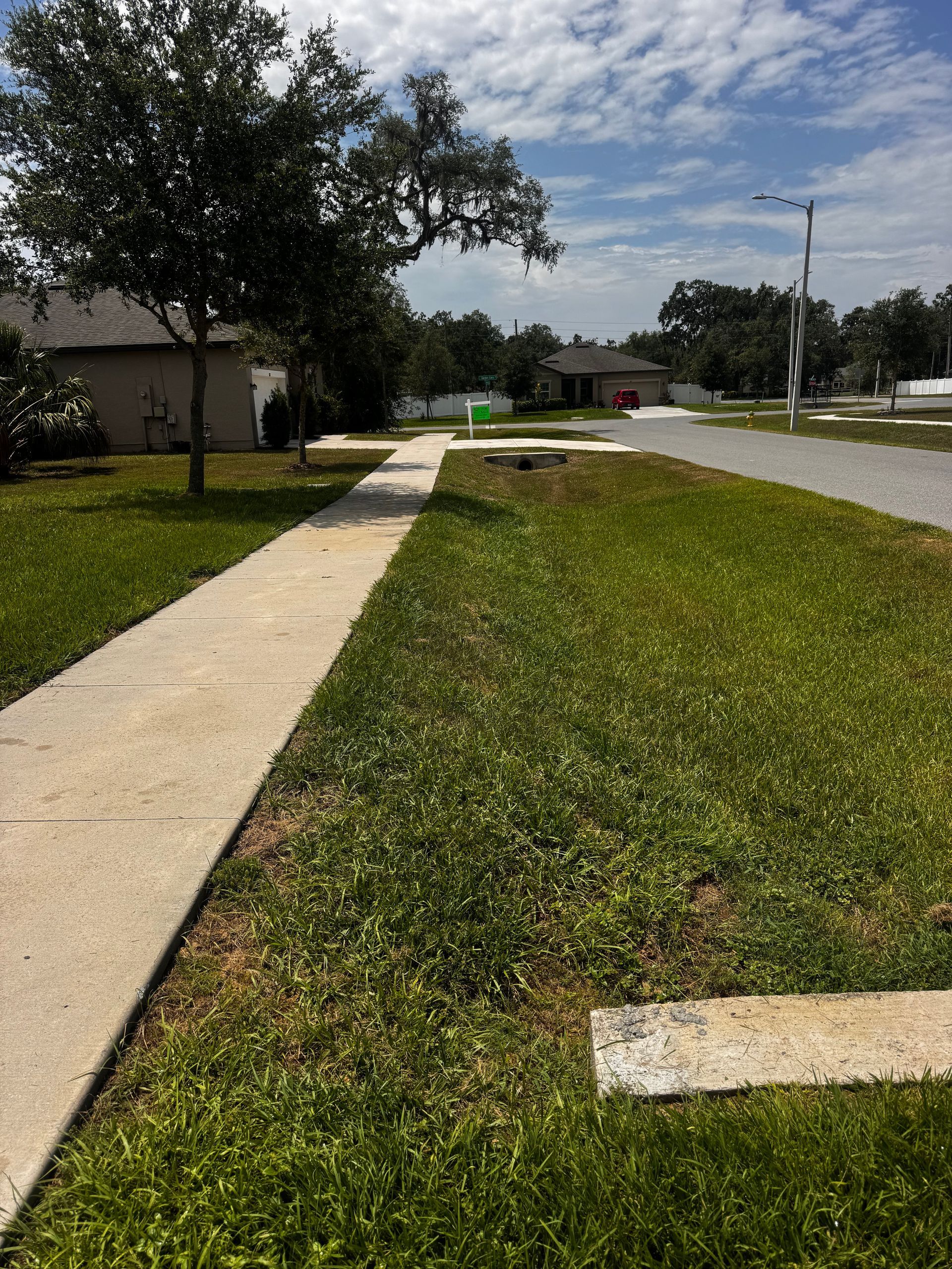 Sidewalk in a residential neighborhood with grass and trees under a cloudy blue sky.