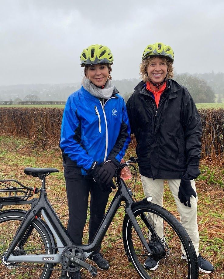 Two cyclists in helmets standing with a road bike on a misty rural path, smiling at the camera.