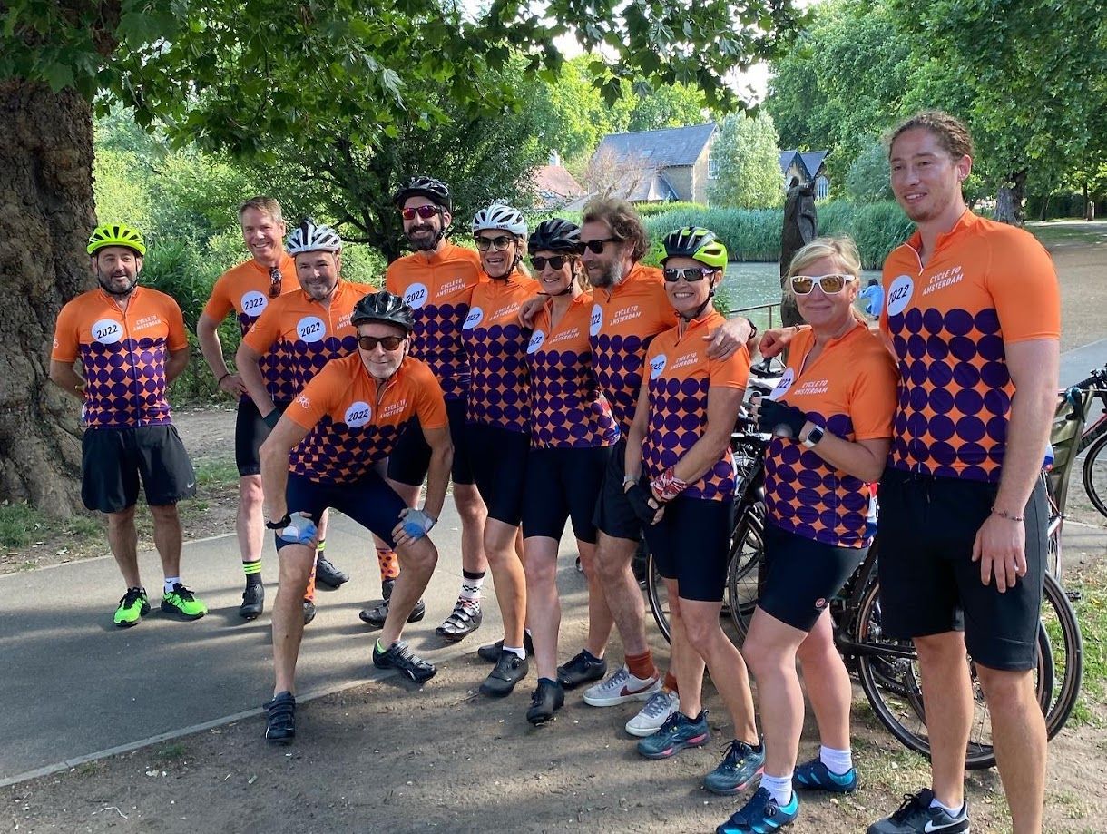 Cyclists in orange and purple jerseys posing together on a tree-lined road, one crouching in front.