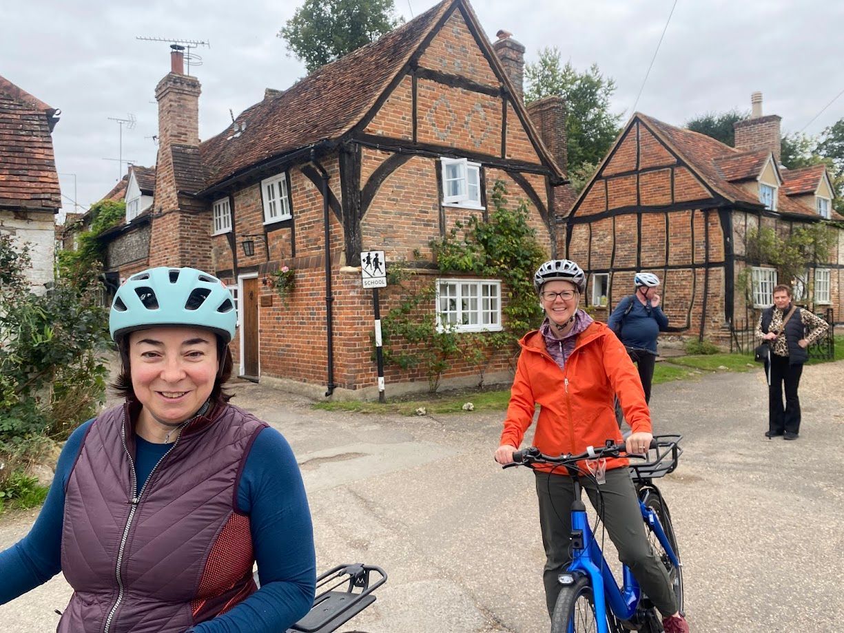 Cyclists on a village street beside brick-and-timber cottages, one smiling woman in a blue helmet foreground.