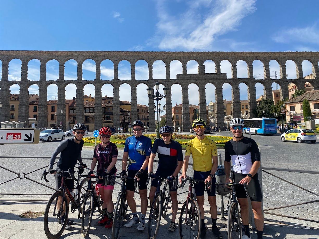 Five cyclists posing with bikes in front of a large stone aqueduct under a blue sky