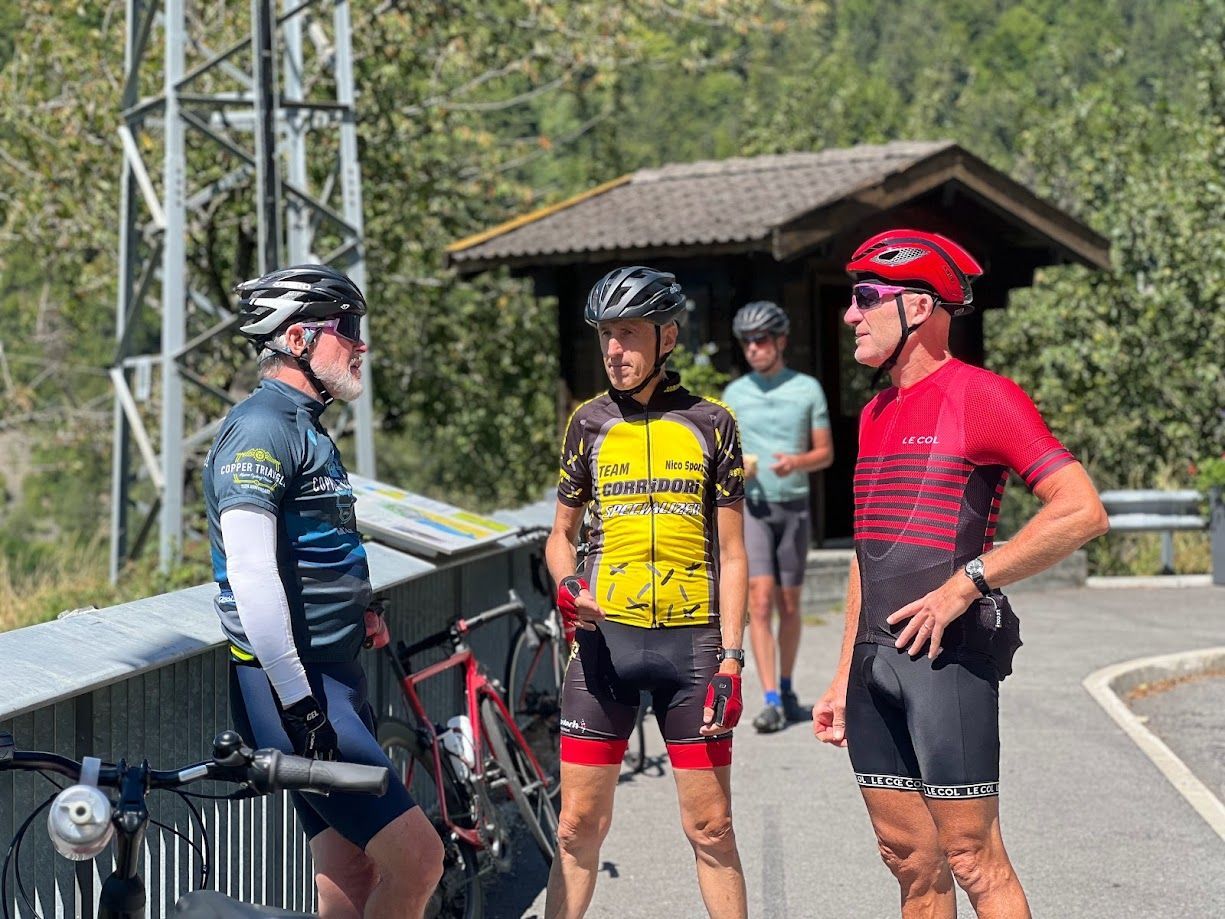 Three cyclists in helmets pause by a road overlooking the mountains.