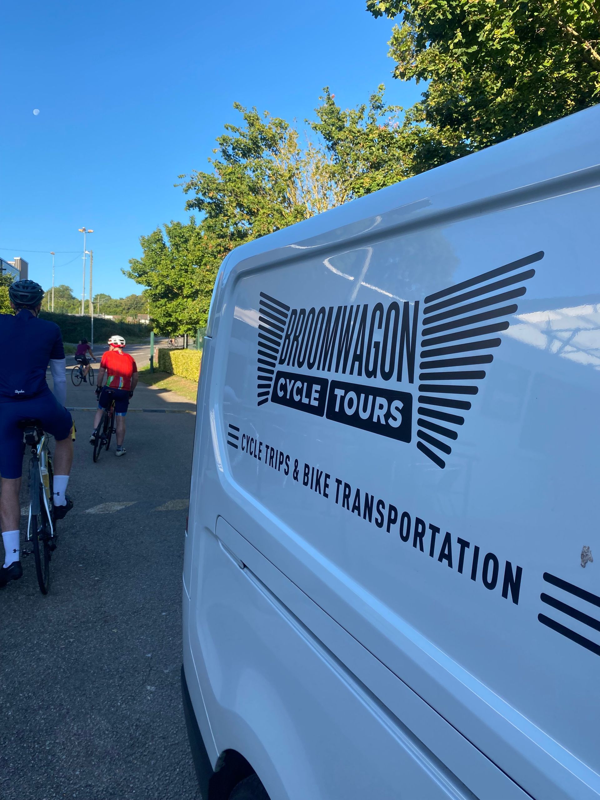 Tour van with logo on roadside near cyclists under a clear blue sky