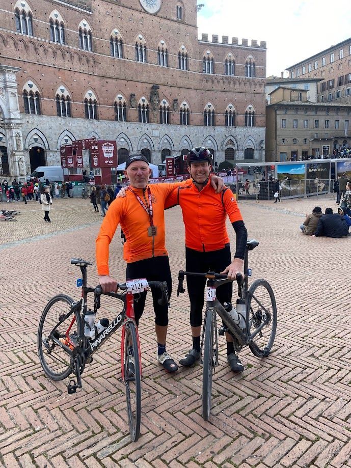 Two cyclists in bright orange jackets standing with bikes in a cobblestone plaza.