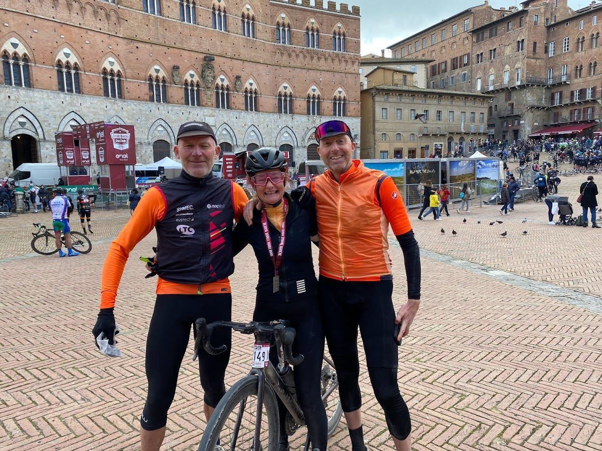Three cyclists in orange-and-black gear posing with bikes in a cobblestone city square