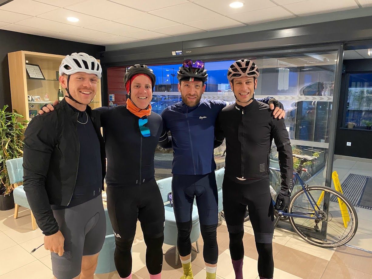 Four cyclists pose indoors in helmets and black gear, standing beside a bike near glass doors.