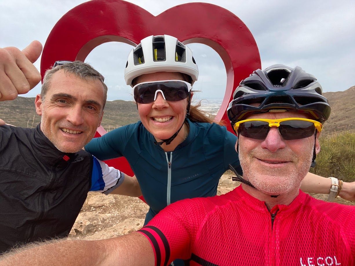 Three cyclists smiling for a selfie with a red heart-shaped sculpture in the background.