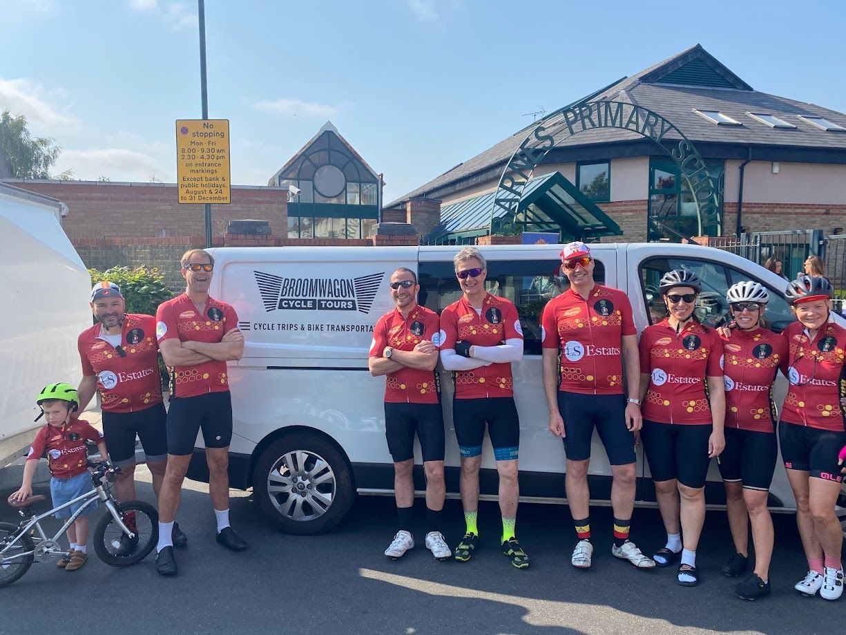 Cyclists in red jerseys posing beside a white van in a parking lot outside a building.