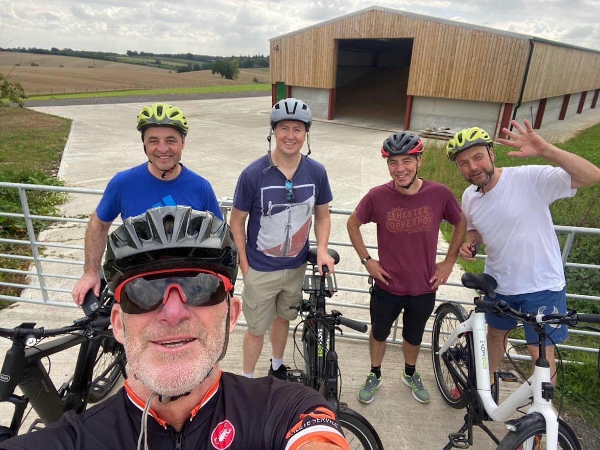 Five cyclists posing with bikes on a rural path beside a covered bridge, one taking a selfie