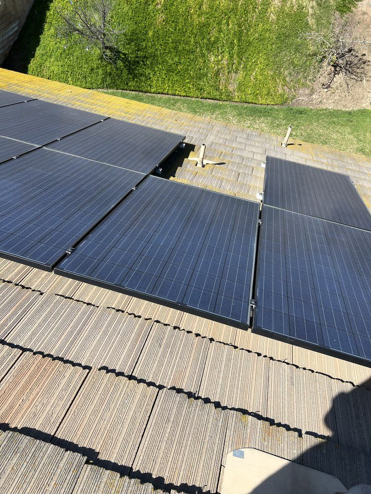 An elevated view of dark solar panels installed on a residential textured tile roof, with a grassy hill visible above.