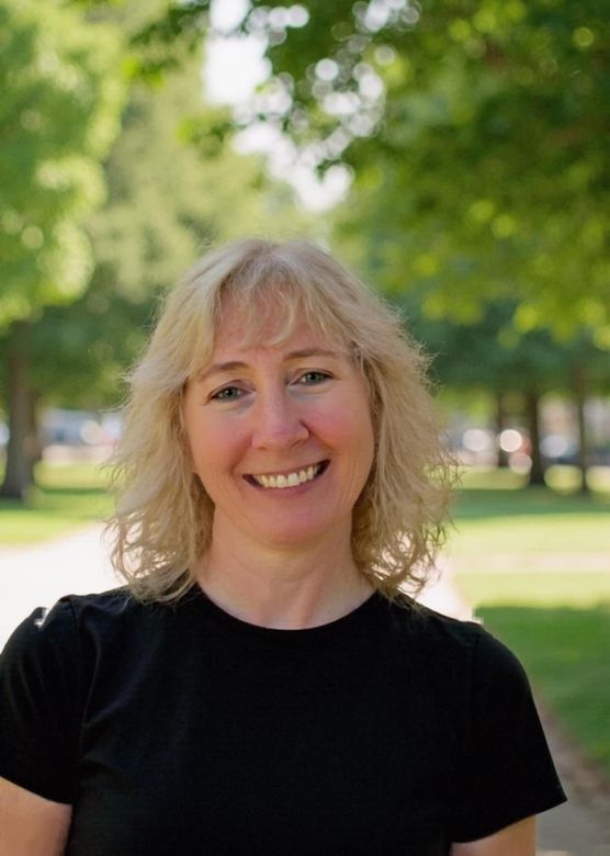 Woman with blonde hair smiling outdoors in front of trees. She wears a black shirt.