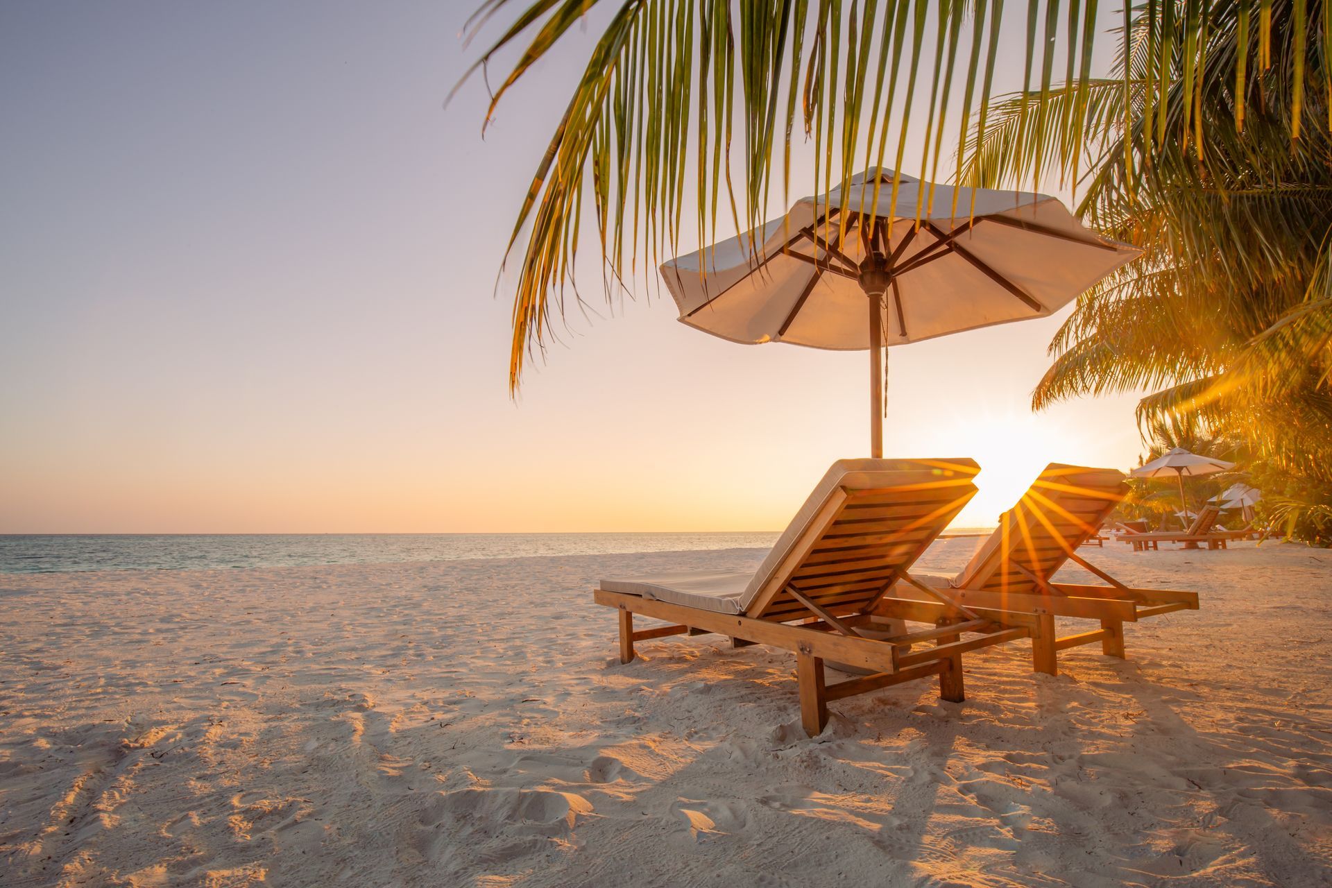 Two lounge chairs under an umbrella on a beach at sunset.