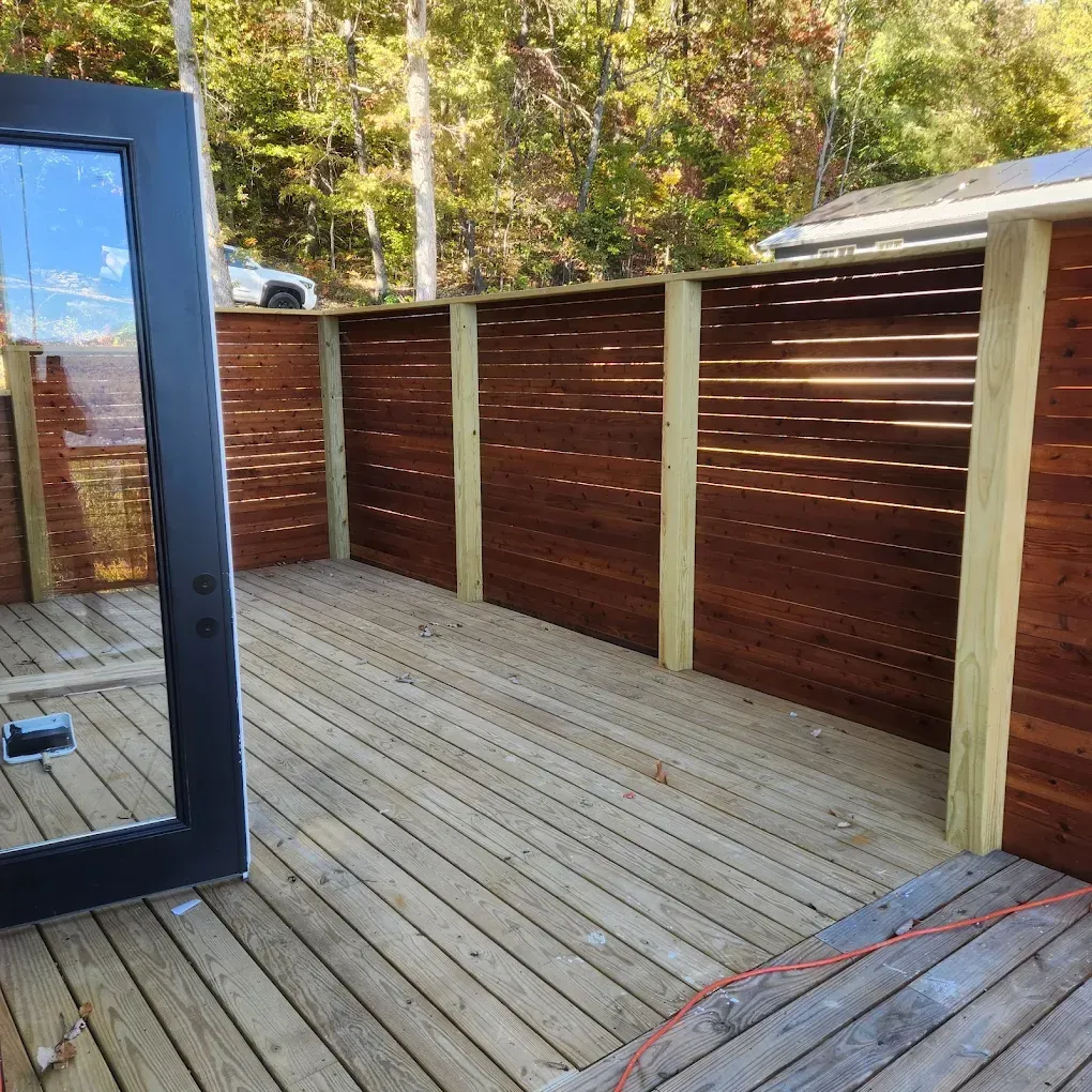 Wooden deck with red-brown privacy fence and glass door on the left, overlooking trees in daylight