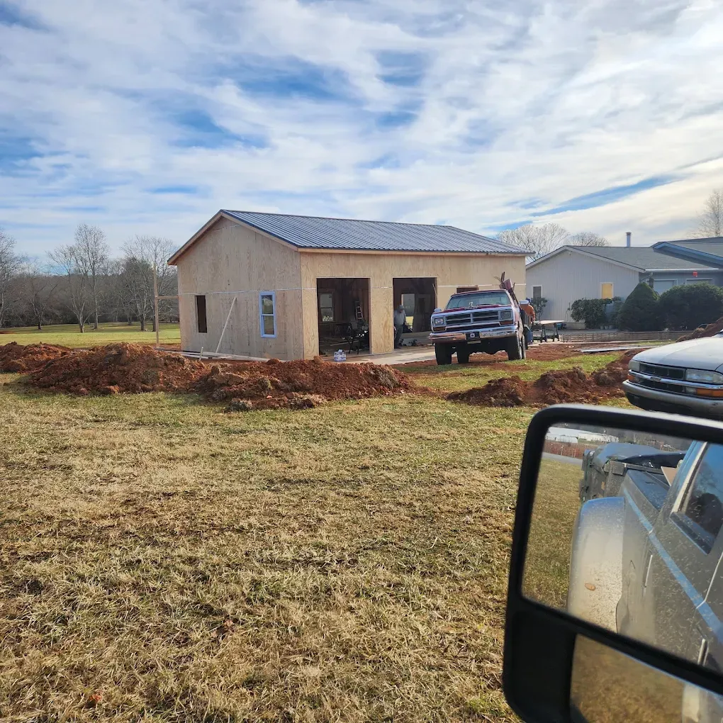 Small beige house under construction with a dark roof, mulch piles, and a pickup truck in the yard