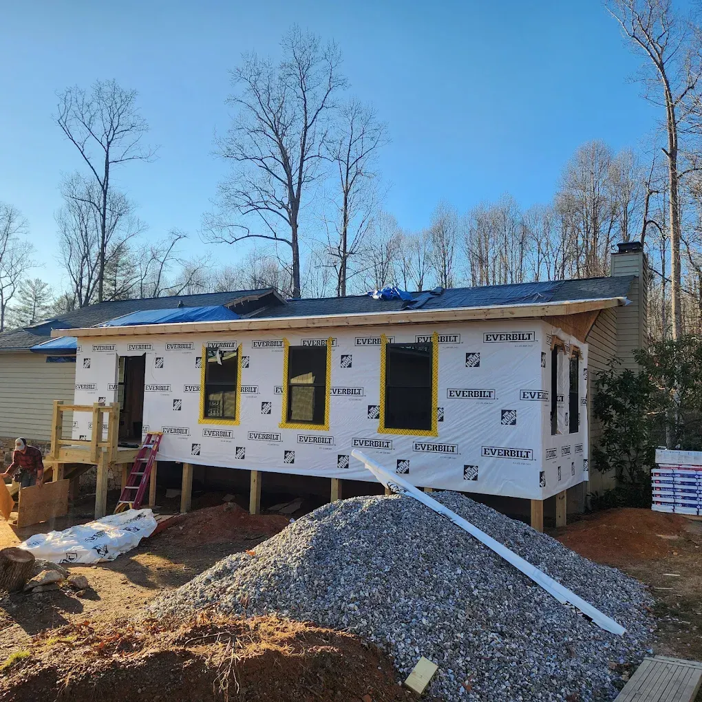 Small house under construction with white wrap, yellow-trimmed windows, and gravel piled in front outdoors.