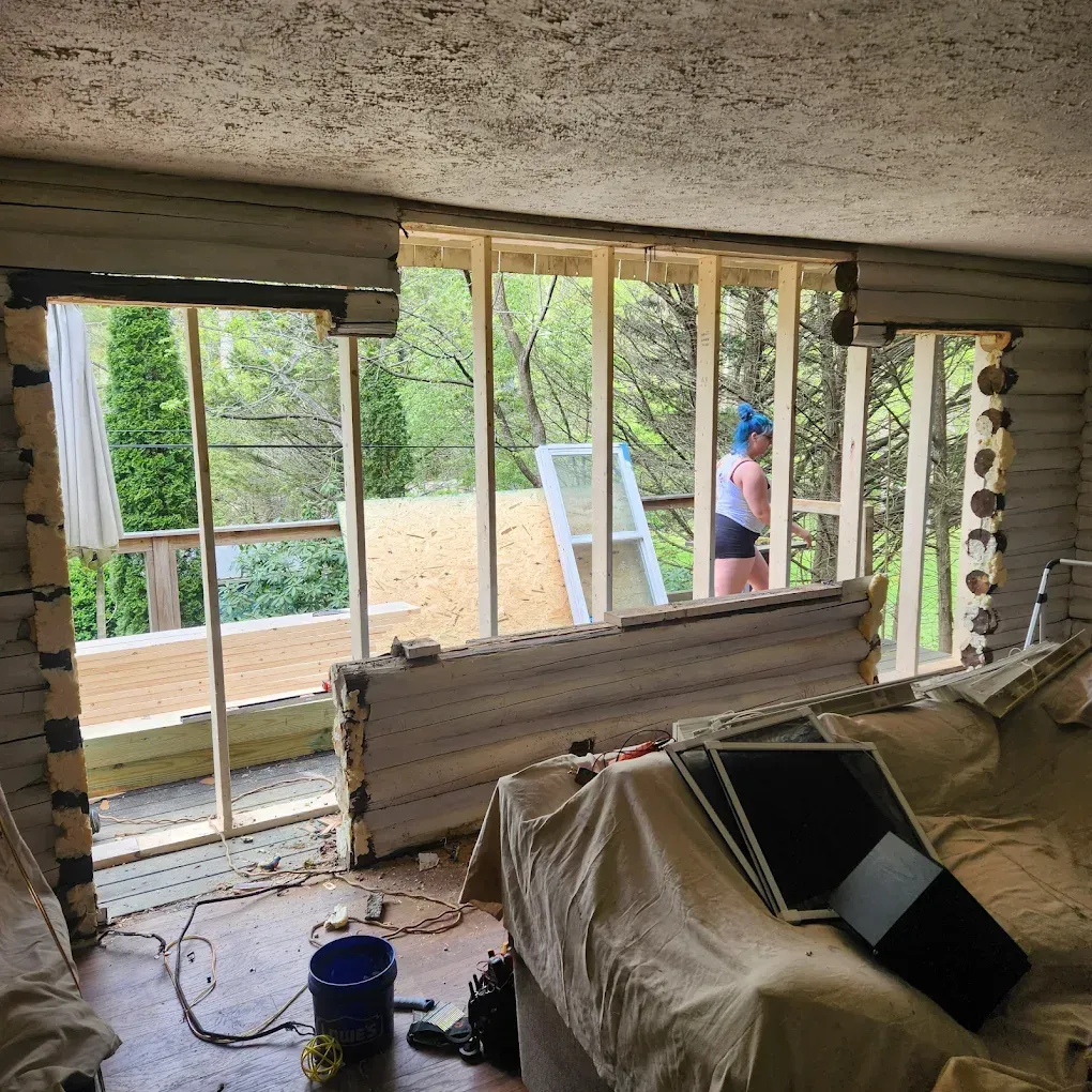 Damaged room with broken wall openings, exposed framing, and construction materials, looking out to a deck and yard.