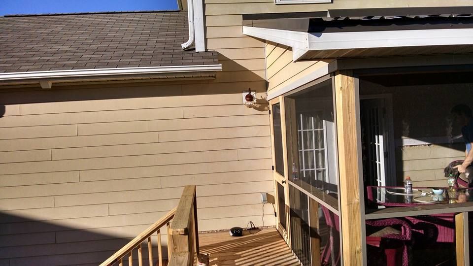 Tan-sided house deck and porch with railing, windows, and a roofline under a blue sky