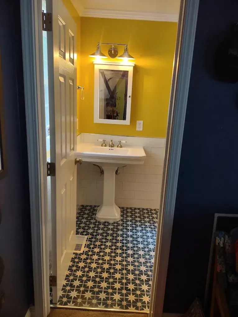 Small bathroom with yellow walls, white pedestal sink, mirror, and patterned black-and-white tile floor.