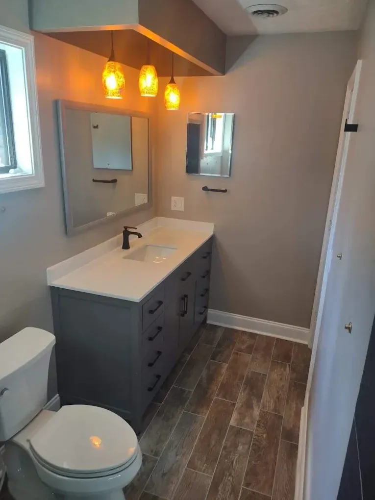 Small modern bathroom with gray vanity, white sink, lit pendant lights, and wood-look floor.
