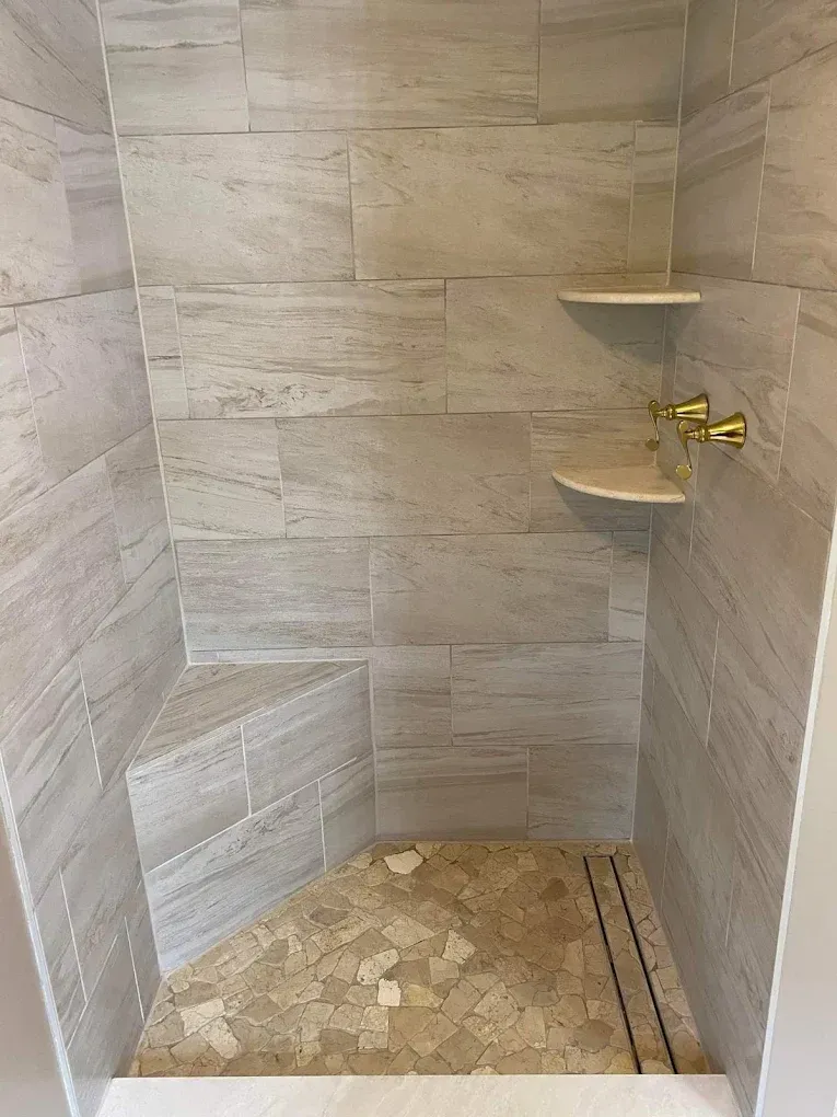 Empty tiled shower stall with two corner shelves and a brass shower fixture, unfinished floor.