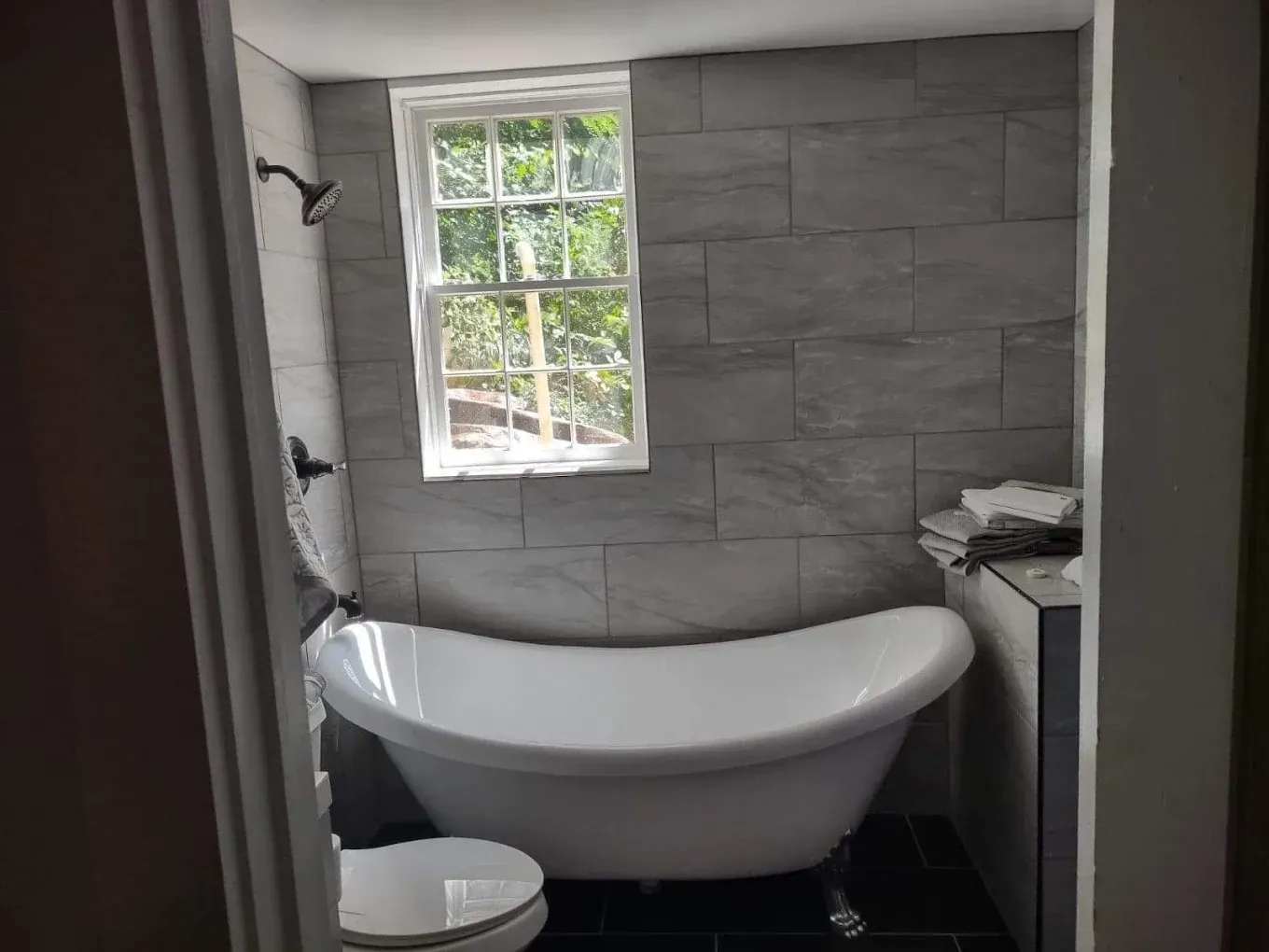 Small bathroom with a freestanding white bathtub under a window and gray tiled walls.