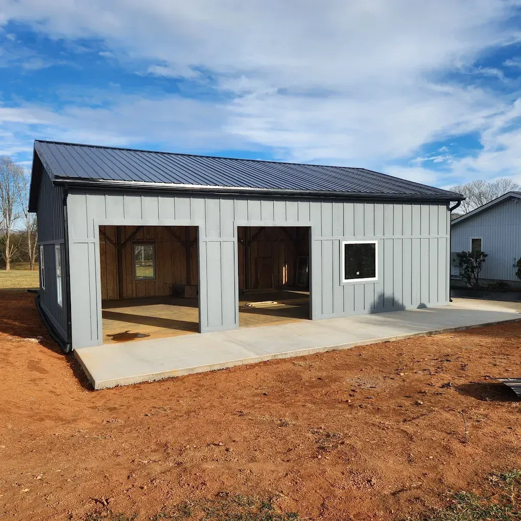 Small gray metal garage or workshop building with open bays, black roof, and dirt foreground under blue sky