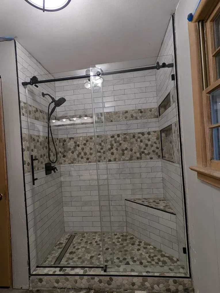 Glass shower stall with white and beige tiled walls, a handheld showerhead, and a bench inside.