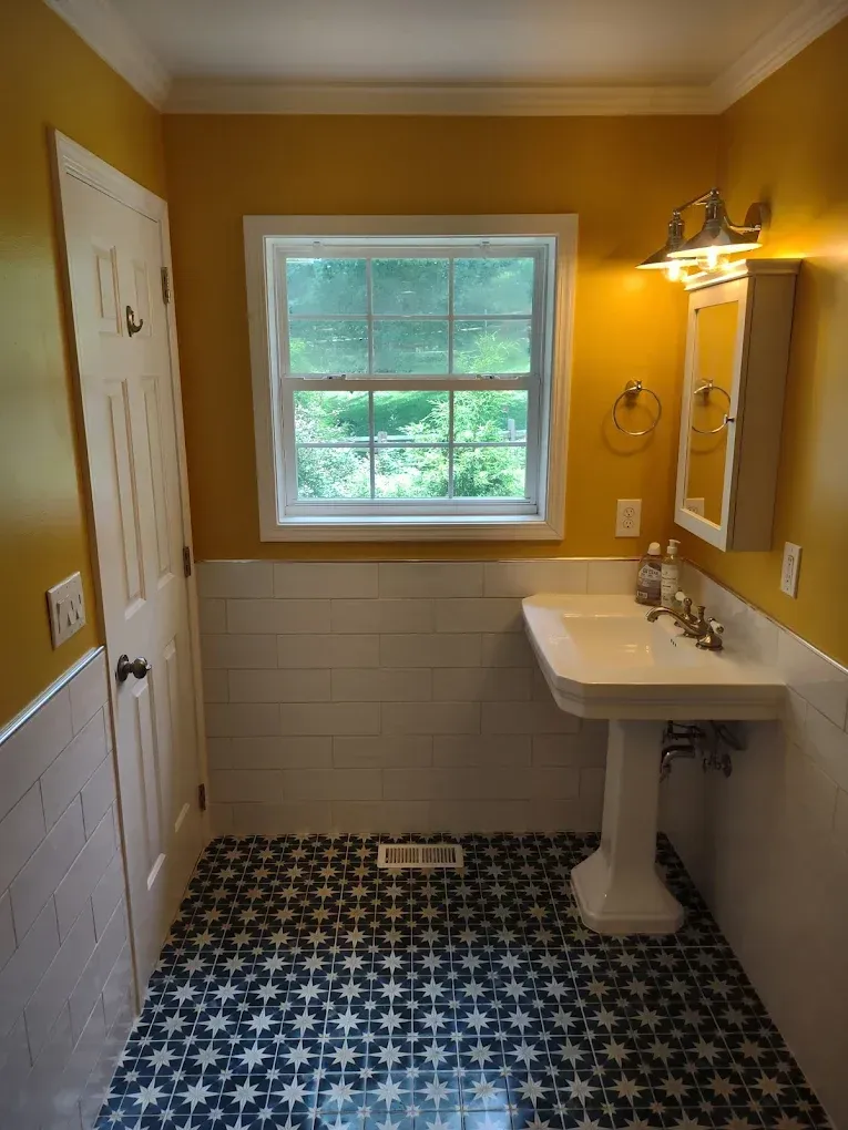 Small bathroom with yellow walls, white pedestal sink, window, and black-and-white patterned tile floor.