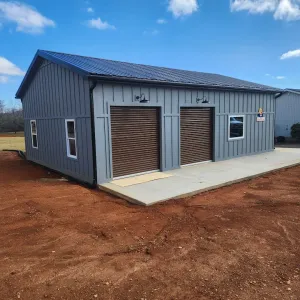 Gray metal garage building with two brown roll-up doors on a concrete slab, in a red dirt lot under a blue sky