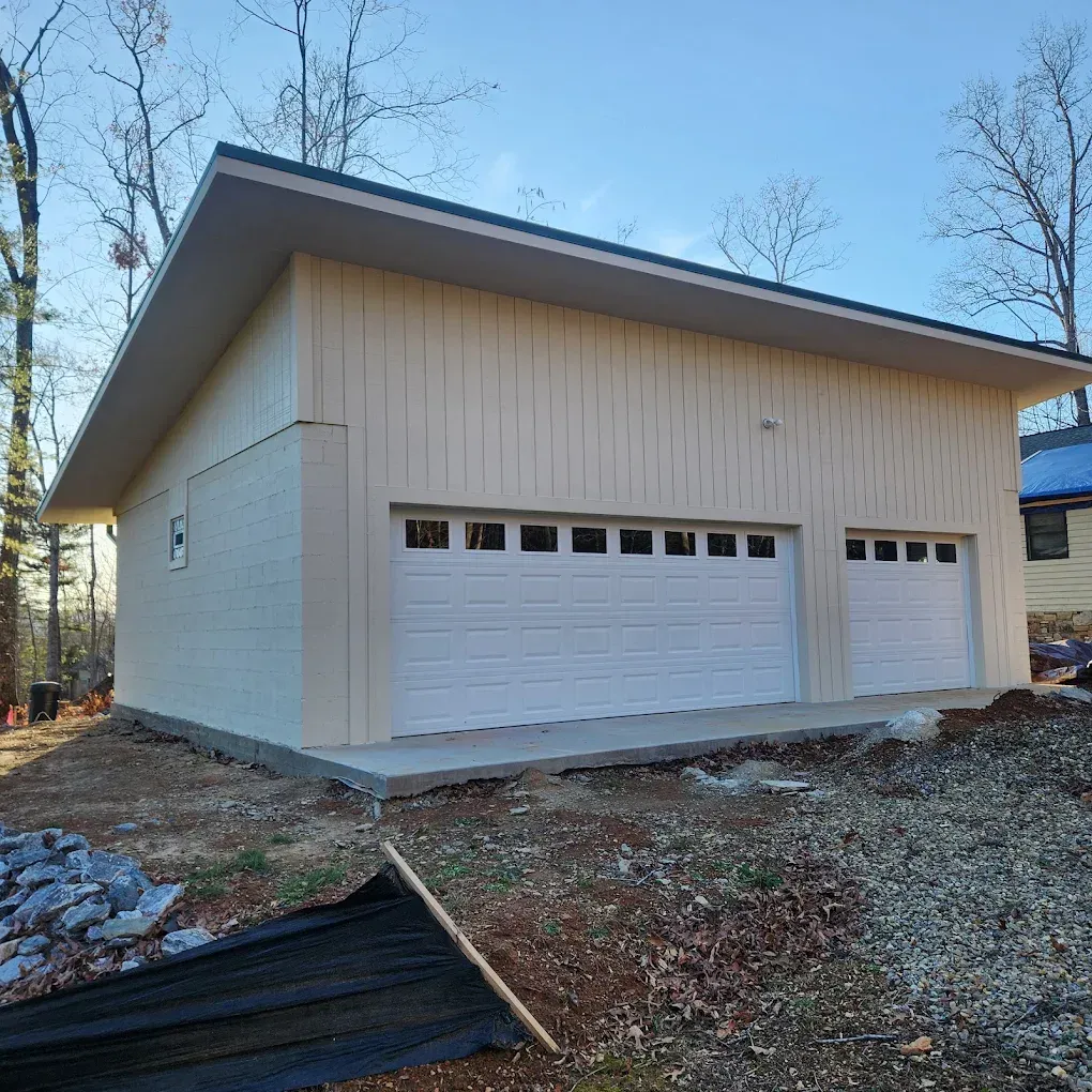White detached garage with two overhead doors on a wooded lot.