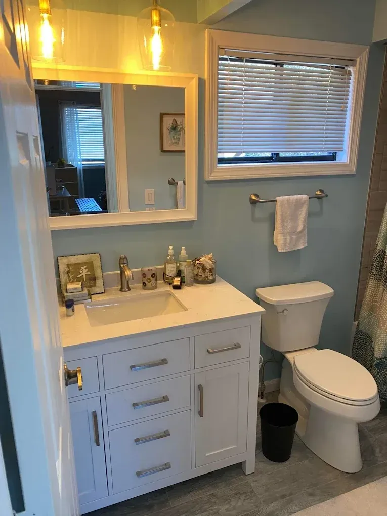 Small bathroom with white vanity, toilet, window blinds, mirror, and warm lights.