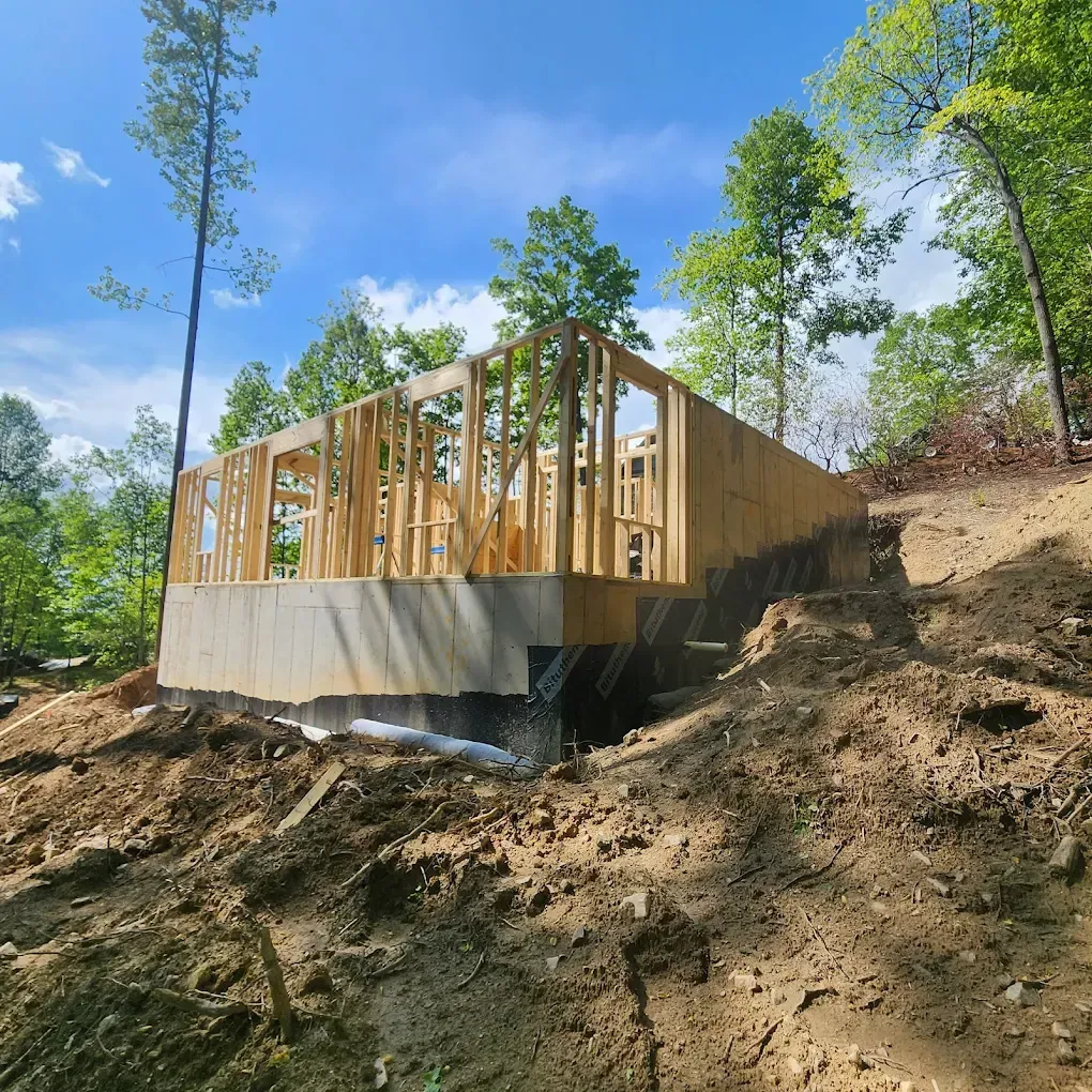 Wood-framed house under construction on a wooded hillside with exposed foundation and dirt slope.