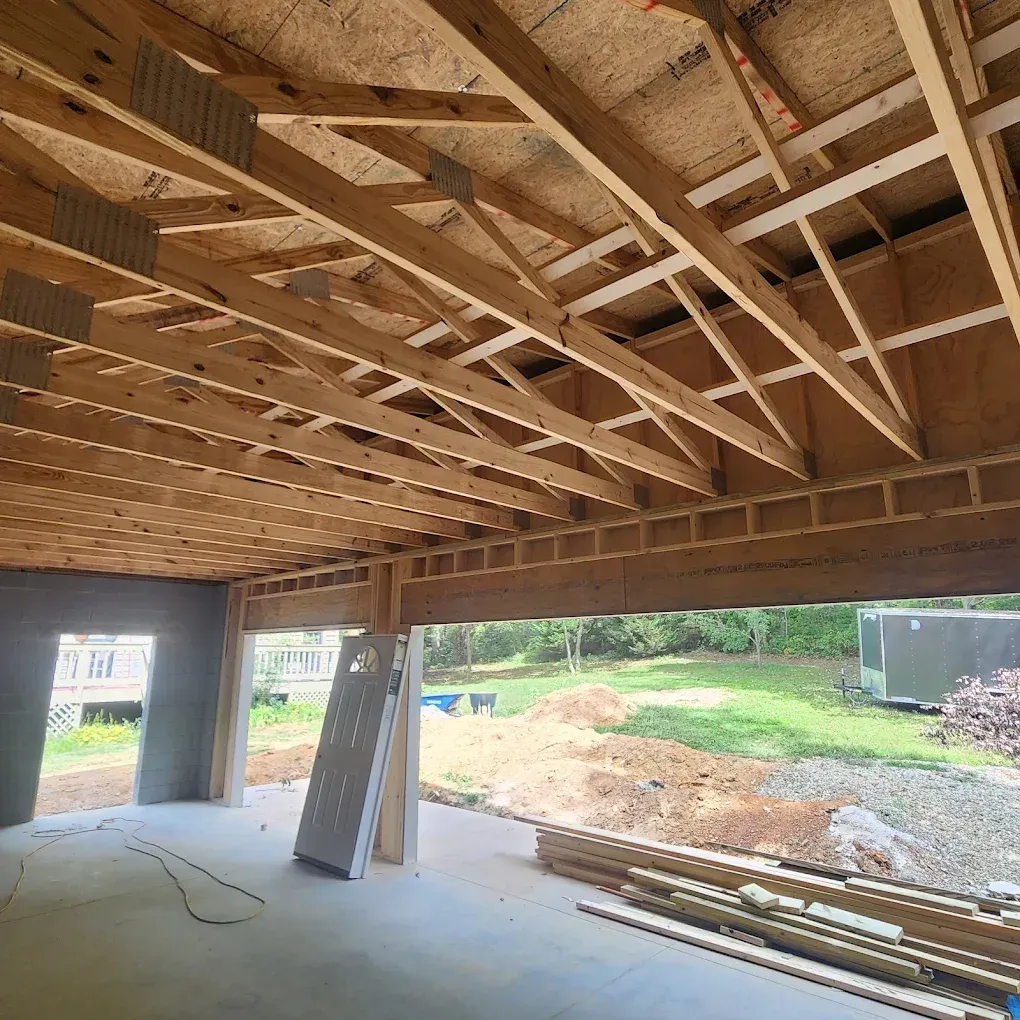 Open deck under construction with exposed wooden roof framing and stacks of lumber on a concrete slab.